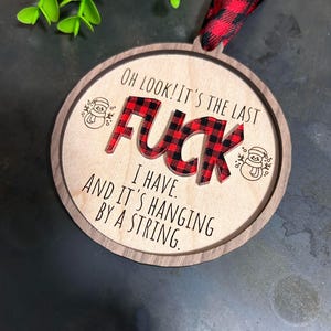 May include: A round wooden Christmas ornament with a red and black plaid design. The ornament says "Oh look! It's the last F**K I have. And it's hanging by a string."