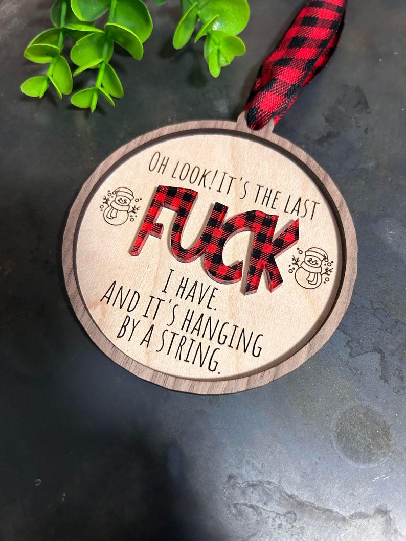 May include: A round wooden Christmas ornament with a red and black plaid design. The ornament says "Oh look! It's the last F**K I have. And it's hanging by a string."