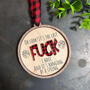 May include: A round wooden Christmas ornament with a red and black buffalo plaid design. The ornament says "OH LOOK! IT'S THE LAST FUCK I HAVE. AND IT'S HANGING BY A STRING."