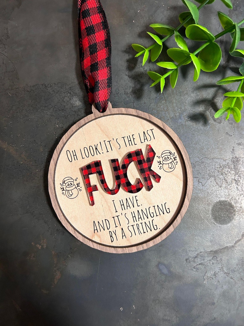May include: A round wooden Christmas ornament with a red and black buffalo plaid design. The ornament says "OH LOOK! IT'S THE LAST FUCK I HAVE. AND IT'S HANGING BY A STRING."