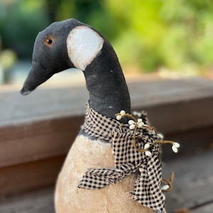 May include: A handmade fabric goose figurine with a black and white head, a brown body, and a black and white checkered ribbon around its neck. The goose is decorated with white berries.