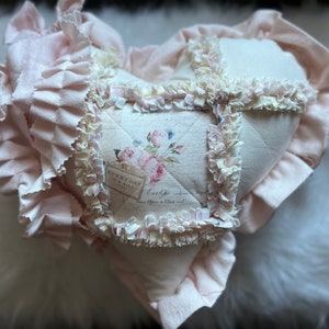 Puede incluir: Un coj&iacute;n de tela en forma de coraz&oacute;n de color rosa p&aacute;lido, con secciones acolchadas y costuras deshilachadas. El panel central muestra un motivo floral con rosas y el texto "EVERYDAY" en un peque&ntilde;o recuadro. Un borde de tela con volantes rodea el coj&iacute;n.