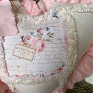Puede incluir: Un coj&iacute;n en forma de coraz&oacute;n con un panel central de color crema que presenta un estampado floral, escritura cursiva y el texto "EVERYDAY IS A GIFT". El coj&iacute;n est&aacute; bordeado por un ribete crema deshilachado y un volante de tela rosa suave.