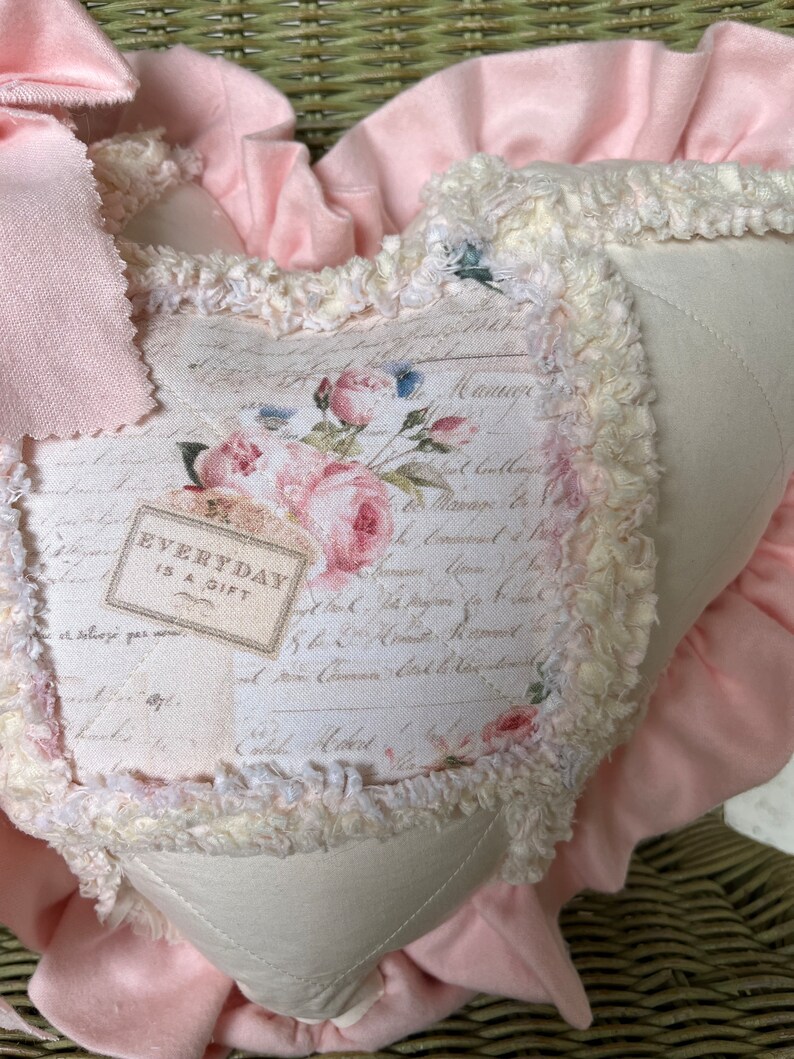 Puede incluir: Un coj&iacute;n en forma de coraz&oacute;n con un panel central de color crema que presenta un estampado floral, escritura cursiva y el texto "EVERYDAY IS A GIFT". El coj&iacute;n est&aacute; bordeado por un ribete crema deshilachado y un volante de tela rosa suave.