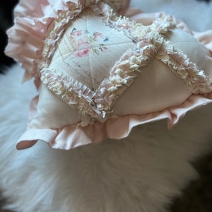 Puede incluir: Un coj&iacute;n en forma de coraz&oacute;n de tela rosa p&aacute;lido con un patr&oacute;n de diamantes acolchado y bordes de tela deshilachada y con volantes en las costuras. El panel central muestra un dise&ntilde;o floral impreso con rosas y texto estilo vintage.