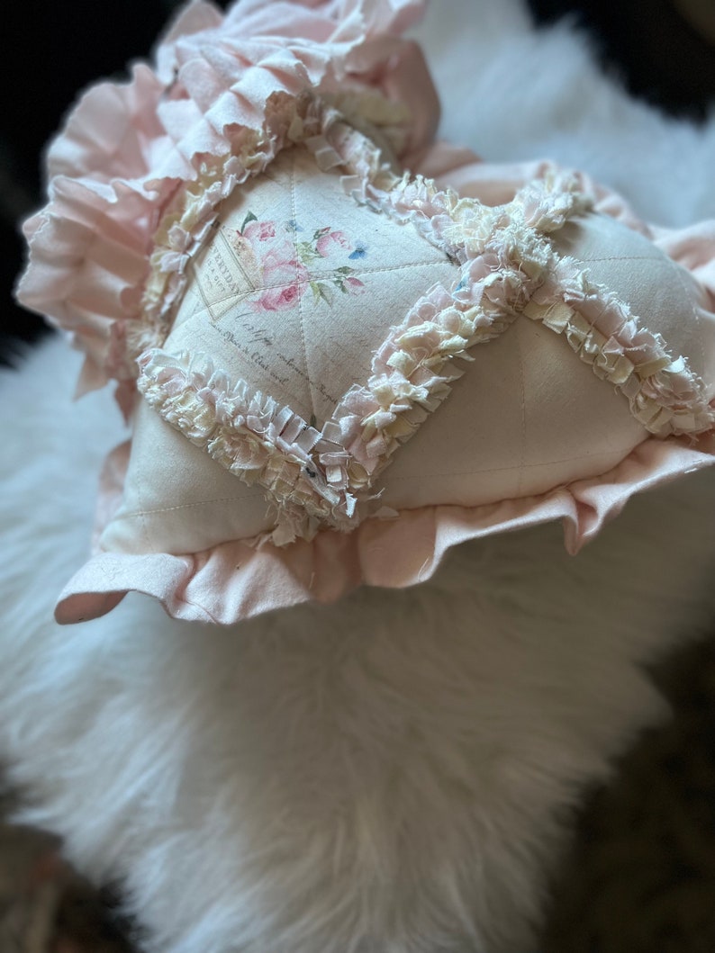 Puede incluir: Un coj&iacute;n en forma de coraz&oacute;n de tela rosa p&aacute;lido con un patr&oacute;n de diamantes acolchado y bordes de tela deshilachada y con volantes en las costuras. El panel central muestra un dise&ntilde;o floral impreso con rosas y texto estilo vintage.