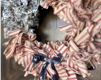 May include: A circular rag wreath constructed from strips of fabric featuring red and white ticking stripes, blue and white floral patterns, and a dark blue ribbon with white stars tied at the bottom. The text House of Bella Rose is visible.