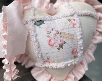 May include: A heart-shaped pillow with a white background and pink ruffles. The pillow is decorated with a floral pattern and the text "Live Life in Full Bloom".