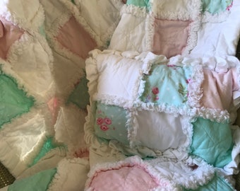 May include: A patchwork quilt with a white, pink, and light blue color scheme. The quilt is made of squares with a ruffled edge and features a floral pattern.