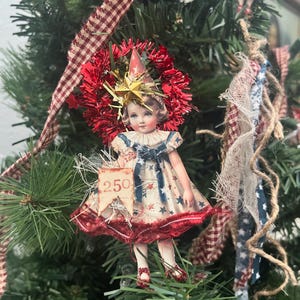 May include: A decorative ornament featuring a vintage-style paper doll illustration wearing a star-patterned dress, accented with a red tinsel halo, a gold star, and a small flag labeled 250. The figure wears red shoes and is displayed on a pine branch.