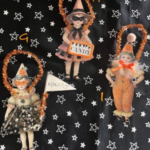 May include: Three vintage-style Halloween decorations on a black background with white stars. The decorations include a witch, a clown, and a figure holding a sign that says "Candy". Each figure is surrounded by a decorative orange ring.