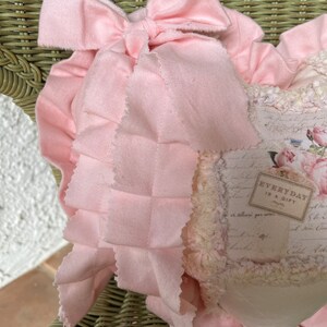 Puede incluir: Un coj&iacute;n decorativo en forma de coraz&oacute;n con un lazo de tela rosa claro y un borde con volantes. El panel central muestra un estampado floral de estilo vintage con texto manuscrito y una etiqueta rectangular que dice EVERYDAY IS A GIFT.
