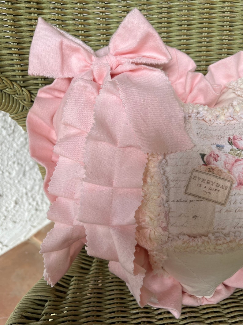 Puede incluir: Un coj&iacute;n decorativo en forma de coraz&oacute;n con un lazo de tela rosa claro y un borde con volantes. El panel central muestra un estampado floral de estilo vintage con texto manuscrito y una etiqueta rectangular que dice EVERYDAY IS A GIFT.