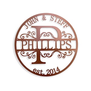 May include: A copper-colored, circular metal sign with the names "JOHN & STEFF" at the top and "PHILLIPS" in the center. The letter "D" and "T" are incorporated into the design, with decorative flourishes. The text "est. 2014" is at the bottom.