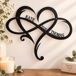Pode incluir: Uma peça de arte de parede em metal preto em forma de símbolo de infinito entrelaçado com um coração. O design apresenta o nome KATE à esquerda, o ano 2026 ao centro e o nome THOMAS no lado direito da estrutura de metal recortada.