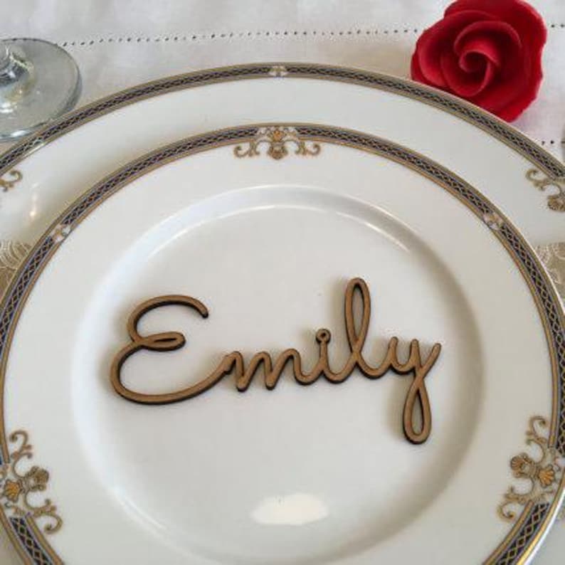 May include: A wooden place card with the name "Emily" written in cursive script. The place card is on a white plate with a gold and black trim.