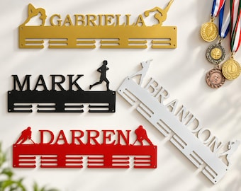 May include: Four metal medal display hangers in gold, black, red, and silver. Each hanger features a personalised name: GABRIELLA, MARK, BRANDON, and DARREN, along with silhouettes of athletes including gymnasts, runners, and hockey players.