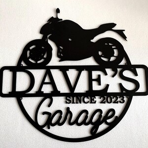 K&ouml;nnte beinhalten: Schwarzes Metallschild mit einer Motorradsilhouette, dem Text "Dave's Garage Since 2023" und einem runden Rand.