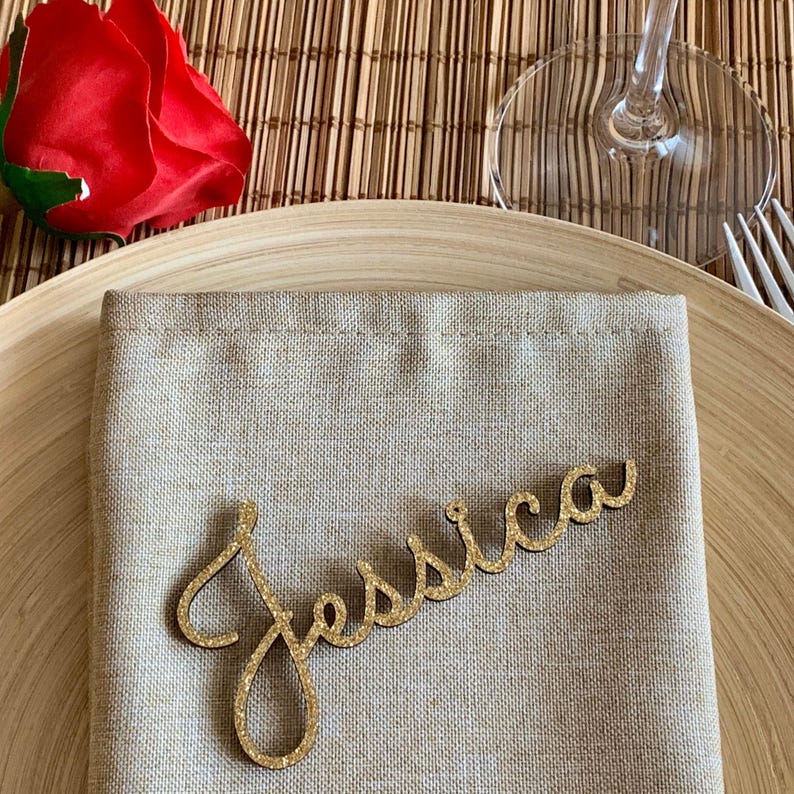 May include: A gold glitter place card with the name "Jessica" written in cursive script.