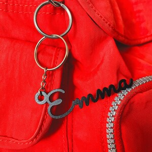 May include: A red backpack with a black metal keychain that says "Emma" in cursive. The keychain has two silver key rings.