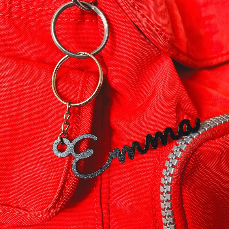 May include: A red backpack with a black metal keychain that says "Emma" in cursive. The keychain has two silver key rings.