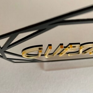 K&ouml;nnte beinhalten: Goldene Buchstaben, die "GUEST" buchstabieren, sind an einem schwarzen geometrischen Rahmen befestigt. Die Buchstaben sind erhaben und haben eine gl&auml;nzende Oberfl&auml;che.