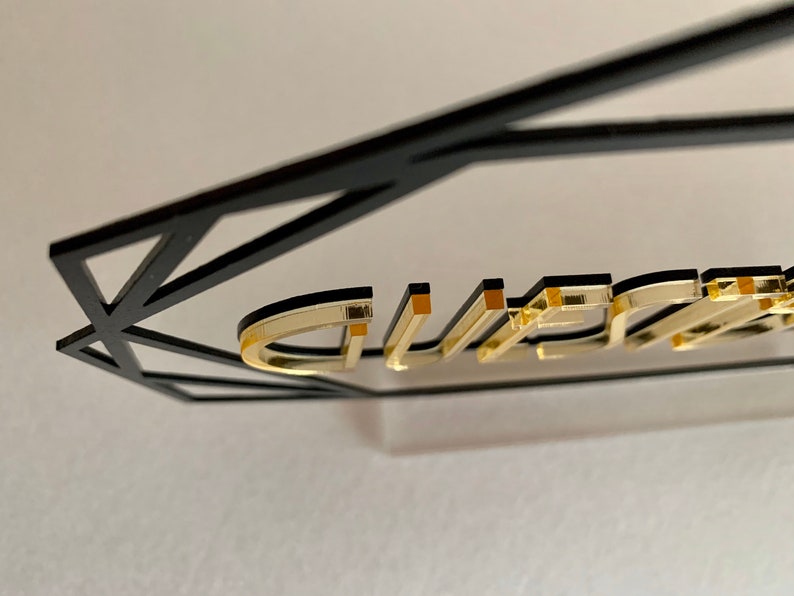 K&ouml;nnte beinhalten: Goldene Buchstaben, die "GUEST" buchstabieren, sind an einem schwarzen geometrischen Rahmen befestigt. Die Buchstaben sind erhaben und haben eine gl&auml;nzende Oberfl&auml;che.