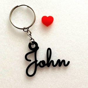 May include: A black metal keychain with the name "John" written in a cursive font. A small red heart charm is attached to the keychain.