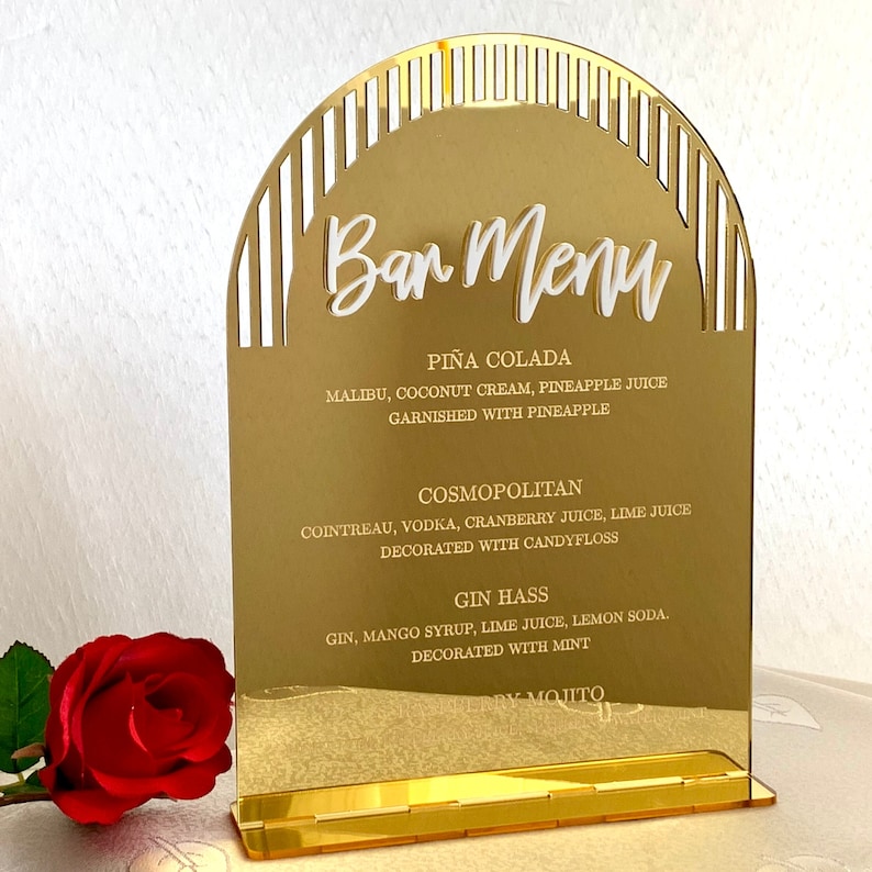 May include: Gold mirrored bar menu sign with a white arch design and white text. The sign lists the drinks: Pi&ntilde;a Colada, Cosmopolitan, Gin Hass, and a blurry text for the last drink.