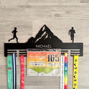May include: Black metal running medal display with a mountain silhouette and the name "MICHAEL". Includes slots for medals and a clear pocket for race bibs. Several colorful race medals and a bib are displayed.