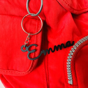 May include: A red backpack with a black metal keychain that says "Emma" hanging from it. The keychain is attached to a silver zipper.