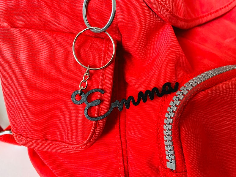 May include: A red backpack with a black metal keychain that says "Emma" hanging from it. The keychain is attached to a silver zipper.