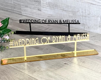 May include: Gold and black acrylic wedding sign with the text "#Wedding of Ryan & Melissa".