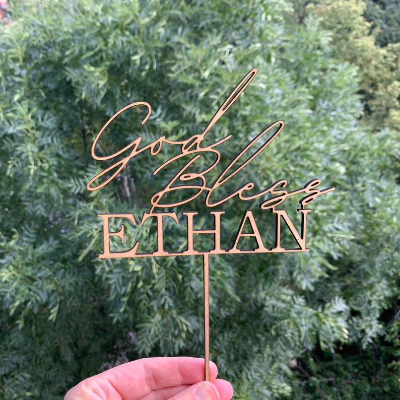 以下が含まれることがあります： 「God Bless Ethan」というテキストが書かれた木製のケーキトッパー。トッパーは茶色で、ケーキに差し込むための棒がついています。
