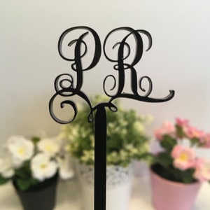 May include: Black metal monogram featuring the letters "PR" in a decorative script font. The monogram is attached to a black stick and is positioned in front of three potted plants.