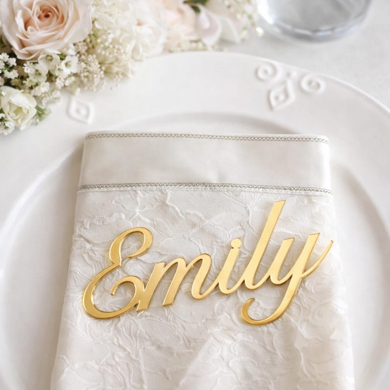 Peut inclure: Une &eacute;tiquette nominative en acrylique dor&eacute; portant le pr&eacute;nom Emily dans une police cursive, pos&eacute;e sur une serviette en tissu blanc &agrave; motif textur&eacute; avec une bordure argent&eacute;e, sur une assiette en c&eacute;ramique blanche.