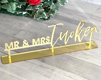 May include: Gold acrylic wedding table sign with the text "MR & MRS Tucker" in a cursive font.