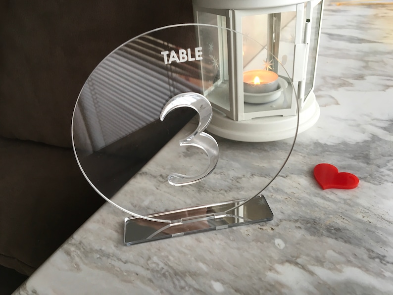 May include: Clear acrylic table number with the word "TABLE" and the number "3" engraved on it. The number is on a clear acrylic stand with a mirrored base.