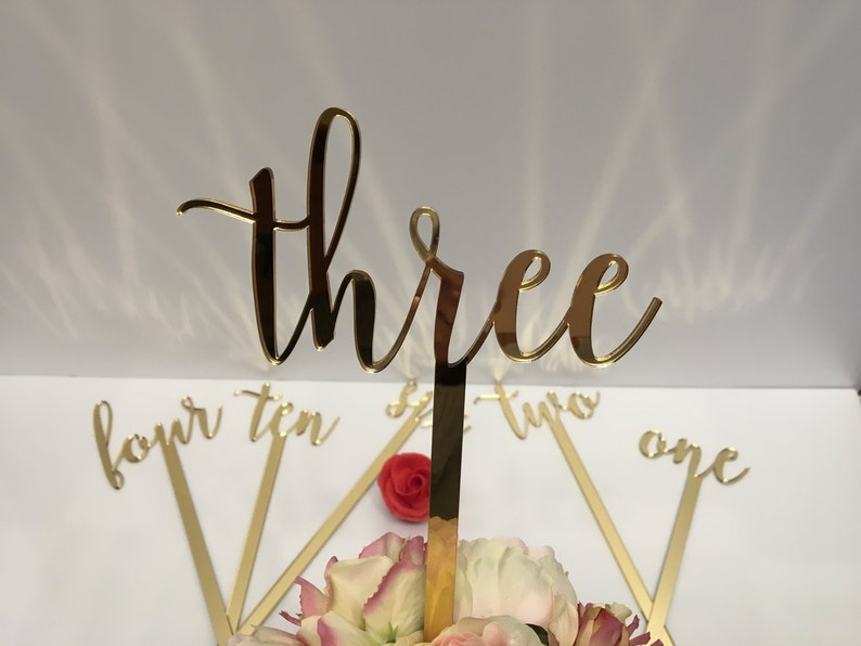 May include: Gold mirror acrylic table numbers with a cursive font. The numbers are on sticks and are arranged in a circle. The numbers include one, two, three, four, and ten.