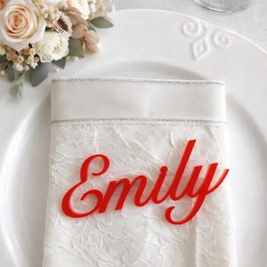 Peut inclure: Une &eacute;tiquette nominative en acrylique rouge vif portant le pr&eacute;nom Emily &eacute;crit en lettres cursives. L'&eacute;tiquette est pos&eacute;e sur une serviette en tissu blanc &agrave; motif floral textur&eacute; avec un liser&eacute; argent&eacute;, sur une assiette en c&eacute;ramique blanche.