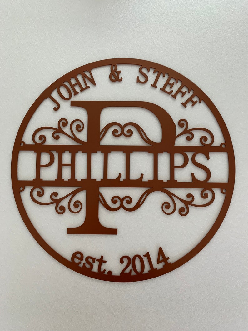 May include: A brown metal family name sign with the names "JOHN & STEFF" above "PHILLIPS" and the year "est. 2014" below. The design features a large letter "P" and decorative flourishes within a circular frame.