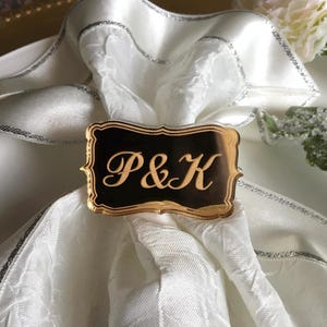 May include: Gold napkin ring with a black rectangular plate featuring the initials "P&K" in gold script.