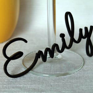 May include: Black acrylic name card with the name "Emily" written in a cursive font. The card is attached to a clear glass wine glass.