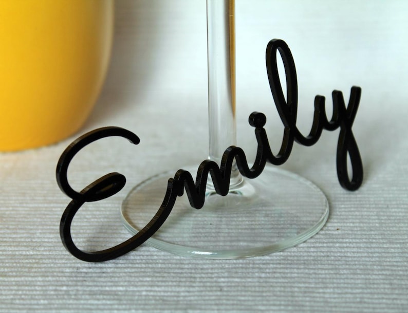 May include: Black acrylic name card with the name "Emily" written in a cursive font. The card is attached to a clear glass wine glass.