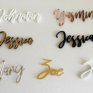 May include: Six acrylic name place cards in various colours, including white, black, gold, silver, and rose gold. The names are written in a cursive font. The names are: Jonathan, Yasmin, Jessica, Mary, Zoe, and Zoe.
