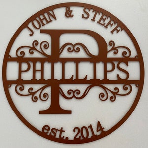 May include: A circular, brown metal sign with the names "JOHN & STEFF" at the top and "PHILLIPS" in the center. The letter "P" is stylized with decorative flourishes. The sign also includes "est. 2014".