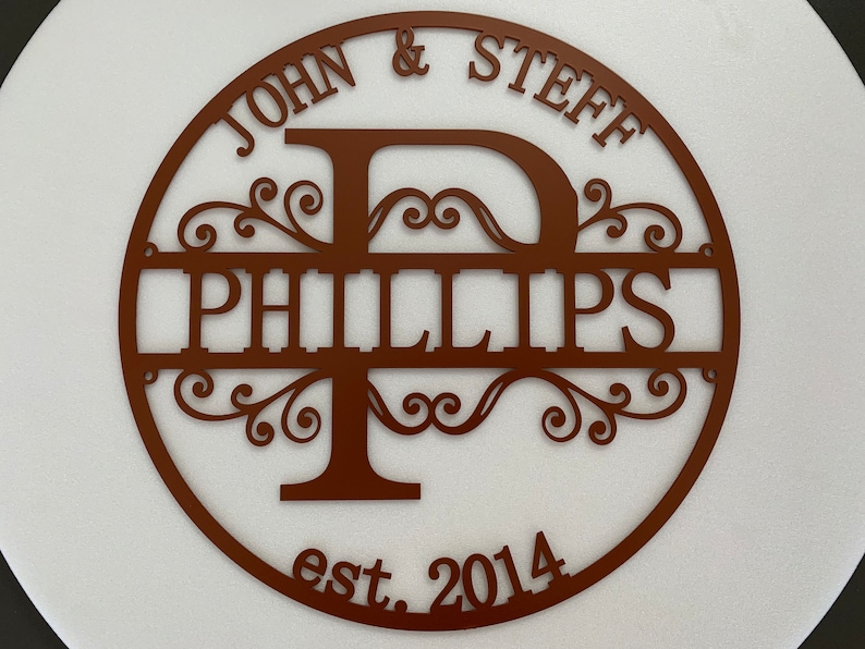 May include: A circular, brown metal sign with the names "JOHN & STEFF" at the top and "PHILLIPS" in the center. The letter "P" is stylized with decorative flourishes. The sign also includes "est. 2014".
