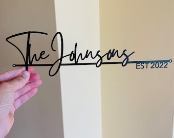 Pode incluir: Placa de metal preta com as palavras "The Johnsons" em fonte cursiva, com "EST 2022" abaixo. A placa é segurada contra uma parede de cor neutra.