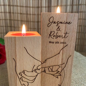 May include: Two wooden candle holders with a couple's names and wedding date engraved on one side. The other side features a design of two hands holding each other. One candle holder has a lit candle inside.