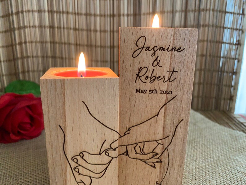May include: Two wooden candle holders with a couple's names and wedding date engraved on one side. The other side features a design of two hands holding each other. One candle holder has a lit candle inside.